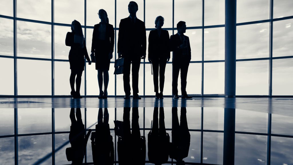 Photo of a group of professionals in a big, open room with floor-to-ceiling windows. They are in business suits, standing confidently.