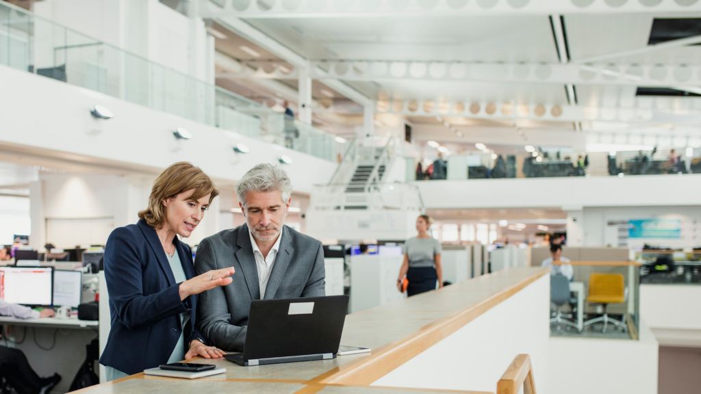 Photo of a man and woman in professional suits talking over a laptop. They are in a big, open office space.