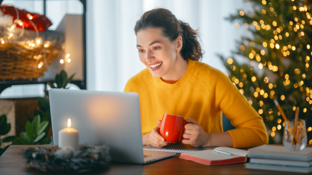 Photo of a woman in a yellow sweater sitting in front of her laptop holding a red mug. Behind her is a christmas tree and twinkling lights.
