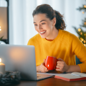 Photo of a woman in a yellow sweater sitting in front of her laptop holding a red mug. Behind her is a christmas tree and twinkling lights.