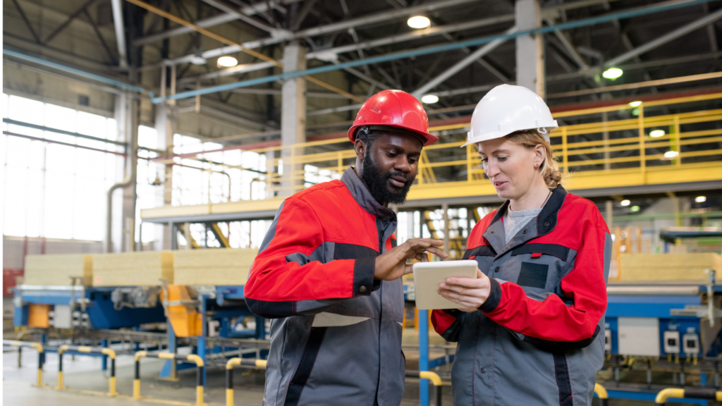 A Black man and white woman working on a tablet in front of manufacturing machinery.