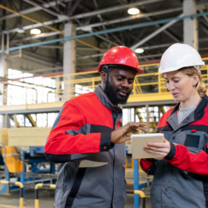A Black man and white woman working on a tablet in front of manufacturing machinery.