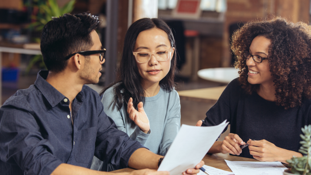 An Asian woman and man sit at a table with a Black woman discussing a resume.